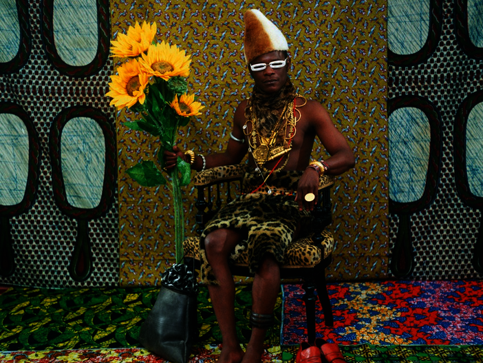 Foto eines Schwarzen Mannes in bunter Kleidung, auf einem Stuhl sitzend, er hat einen Strauß Sonnenblumen in der rechten Hand